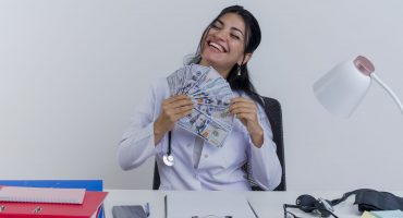 joyful young female doctor wearing medical robe and stethoscope sitting at desk with medical tools holding money with closed eyes isolated on white background